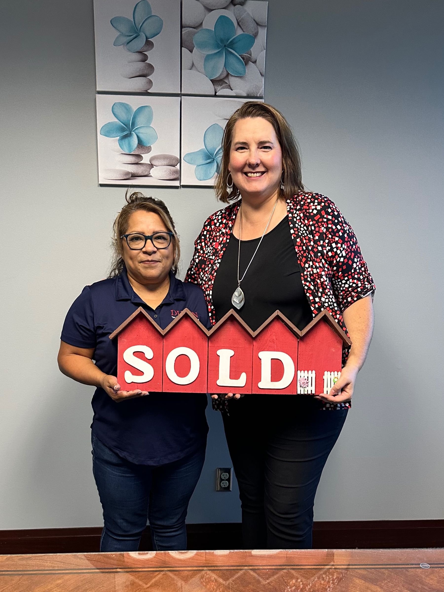 Lisa Collio and a client standing together indoors, holding a red house-shaped sign reading "Sold."